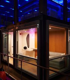 Night view through glass of a cozy modern lounge pod with gray sofa, wood-paneled walls, vanity mirror and blue LED exterior lighting.