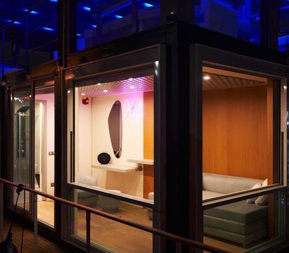 Night view through glass of a cozy modern lounge pod with gray sofa, wood-paneled walls, vanity mirror and blue LED exterior lighting.