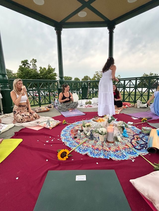 Outdoor wellness circle in a park gazebo — people on mats and cushions gathered around a colorful boho mandala cloth with candles, crystals, a sunflower and ritual items, green railings and lake trees visible beyond.