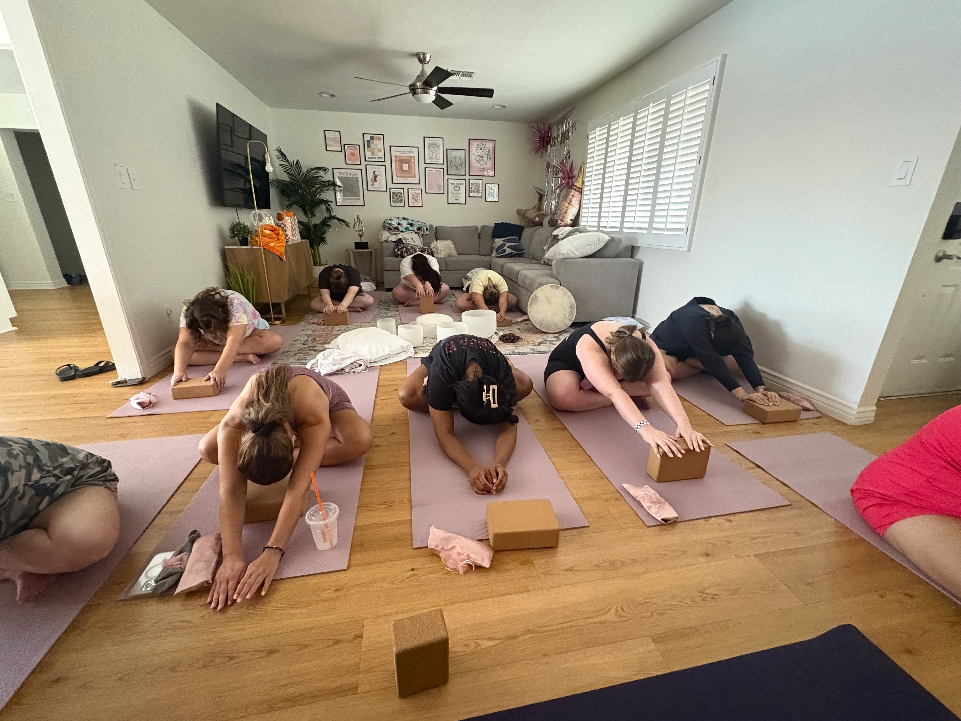 Bright living-room yoga session with participants on pink mats using cork blocks in child's pose on hardwood floor, cozy couch, gallery wall and plants in the background — home group yoga class.