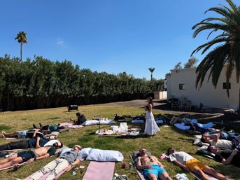 Outdoor sound bath and guided meditation circle on a sunny lawn with palm trees, people lying on mats in a ring around an instructor and sound bowls under a clear blue sky