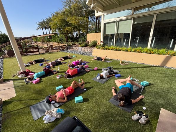 Sunny outdoor yoga class on a landscaped lawn with participants lying on mats and foam blocks near a glass-walled studio and palm trees, outdoor fitness and relaxation scene