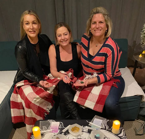 Three smiling women seated on a couch, wrapped in red-and-white checkered blankets, holding colorful crystals over a candlelit crystal-healing setup with bowls of stones on a low table.