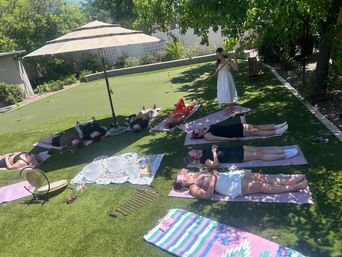 Outdoor sound-bath on a sunny backyard putting green: people lie on yoga mats under trees and an umbrella while a facilitator plays a long tone instrument; crystal singing bowls, gong and chimes arranged on the grass.