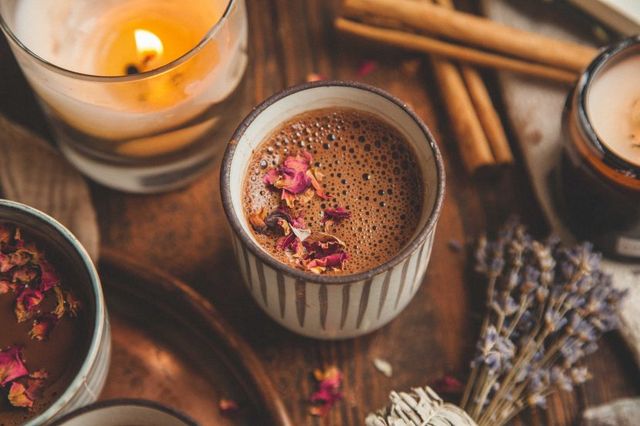 Frothy spiced hot chocolate in a striped ceramic cup garnished with dried rose petals on a rustic wooden tray, surrounded by lit candles, cinnamon sticks and dried lavender for a cozy autumn vibe.