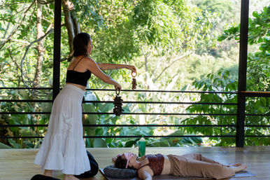 Woman leading an outdoor yoga/meditation sound-healing session on a wooden deck at a tropical retreat, holding a shell rattle over a blindfolded participant lying on a mat
