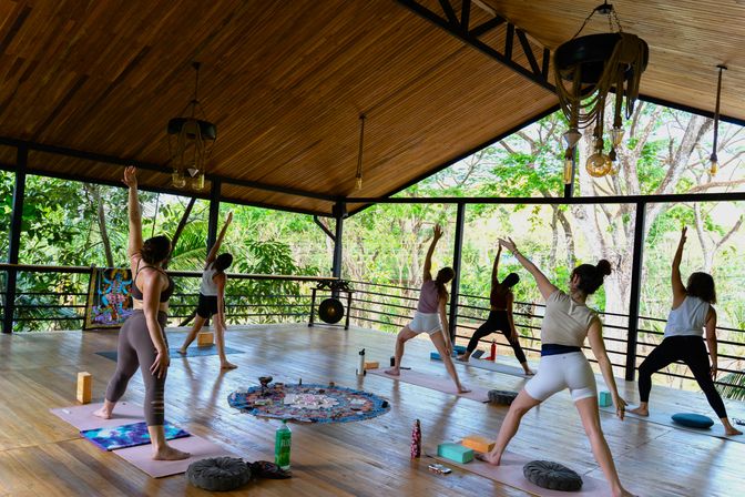 Open-air tropical yoga class on a covered wooden pavilion, students practicing standing warrior poses on mats with lush green trees visible beyond the railings