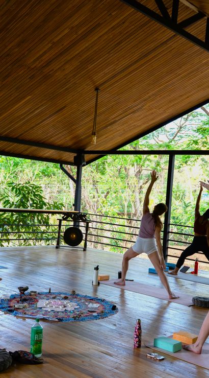 Open-air tropical yoga class on a covered wooden pavilion, students practicing standing warrior poses on mats with lush green trees visible beyond the railings