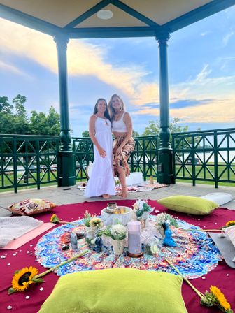 Two women standing barefoot in a lakeside park gazebo at sunset, smiling over a bohemian picnic spread with a colorful mandala cloth, cushions, sunflowers, candles and flowers on a burgundy blanket, lake and trees visible in the background.
