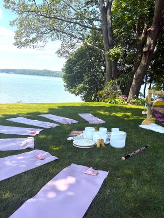 Serene lakeside yoga and sound-healing setup with pink yoga mats arranged on a green lawn under shady trees, crystal singing bowls, drums and a wooden frame beside a calm blue lake.