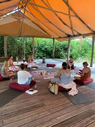 Participants seated on red cushions in a circle inside an open-air wooden pavilion with an orange canvas roof, meditating and sharing around a colorful mandala of crystals in a tropical wellness retreat