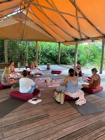 Participants seated on red cushions in a circle inside an open-air wooden pavilion with an orange canvas roof, meditating and sharing around a colorful mandala of crystals in a tropical wellness retreat