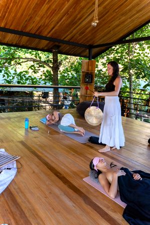 Outdoor sound bath at a tropical wellness retreat: facilitator striking a gong on a wooden open-air deck while participants rest on yoga mats