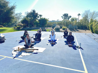Group of people seated on yoga mats for an outdoor class on a blue sports court, surrounded by desert landscaping, palm trees and bright sunny sky.