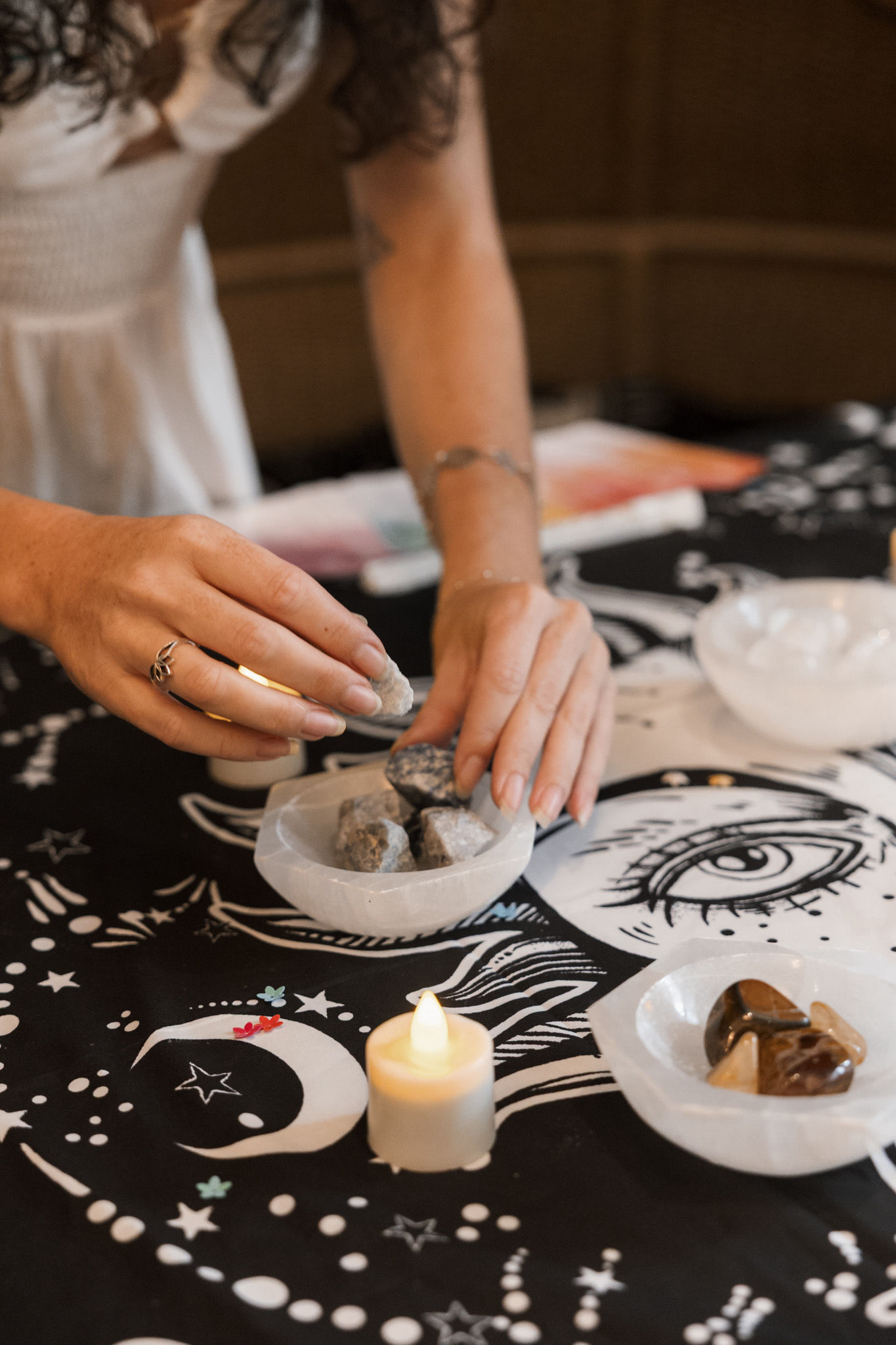 Hands arranging raw crystals in white selenite bowls on a black celestial cloth with a crescent moon, stars and an illustrated eye, a lit tealight candle adding a cozy ritual vibe.