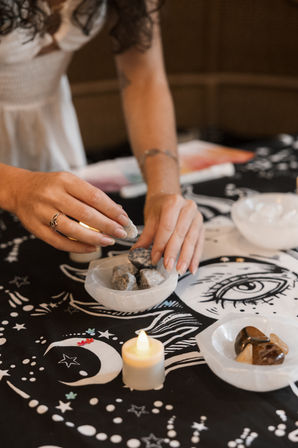 Hands arranging raw crystals in white selenite bowls on a black celestial cloth with a crescent moon, stars and an illustrated eye, a lit tealight candle adding a cozy ritual vibe.