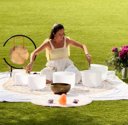 Person seated on a blanket on a sunny grassy lawn leading an outdoor sound bath with white crystal singing bowls, a bronze singing bowl and gong, a Himalayan salt lamp and a bouquet of flowers.
