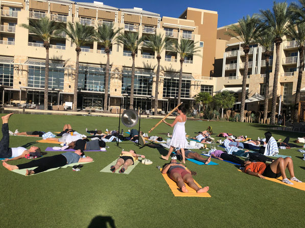 Sunny resort lawn sound-bath: people lying on colorful yoga mats in savasana while a woman in white plays a long wooden wind instrument, framed by palm trees and hotel balconies.