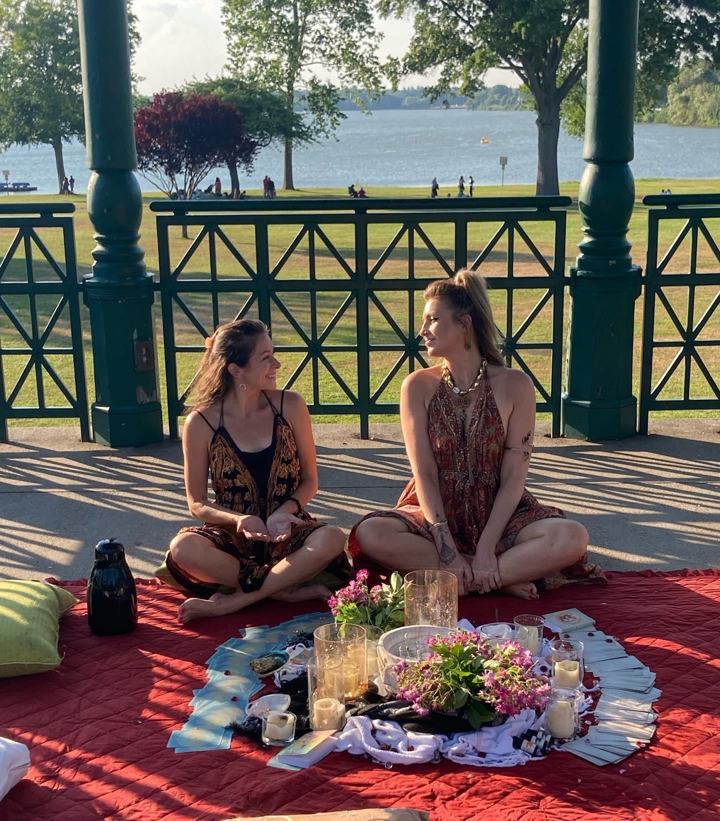 Two friends sitting cross-legged on a red blanket inside a green lakeside bandstand, enjoying a sunny picnic setup of candles, flowers and cards with a grassy park and lake in the background.