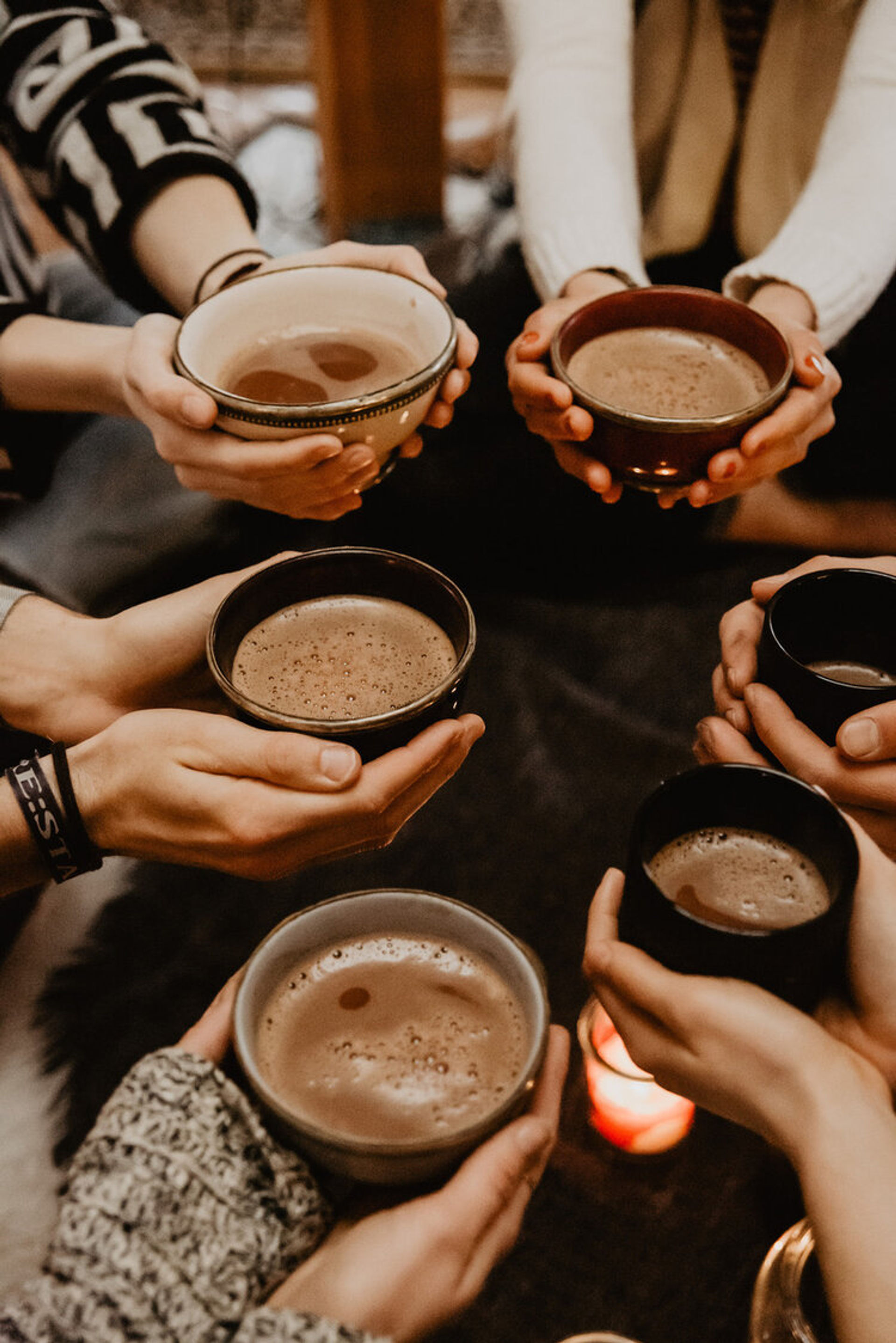 Hands holding ceramic bowls and mugs of hot chocolate in a cozy candlelit indoor gathering — warm drinks shared among friends.