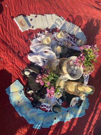 Overhead boho outdoor tarot altar on a red quilt — fanned blue tarot cards encircling lit candles, glass bowls, crystals, white cloth and pink-purple flowers.