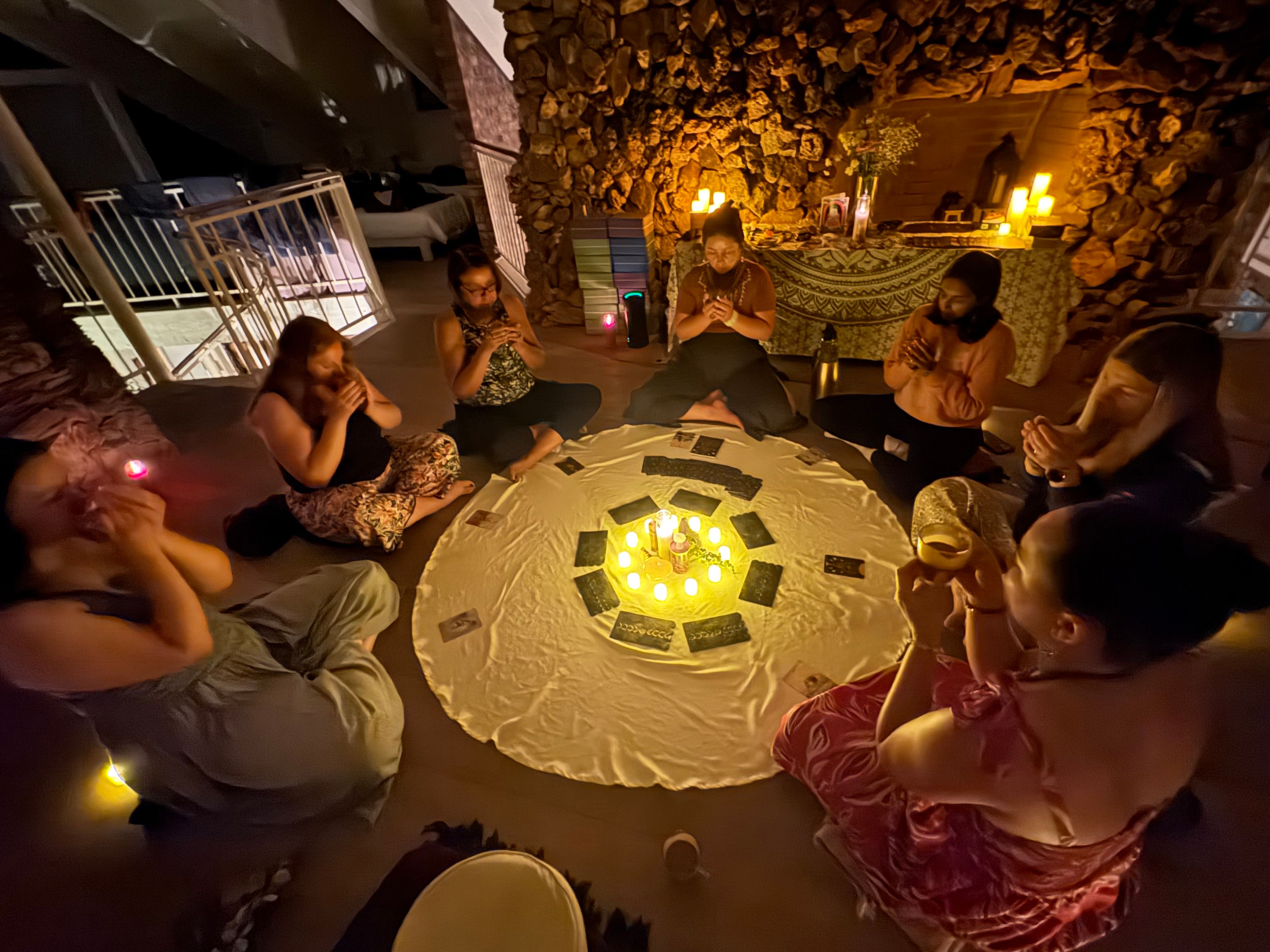 Candlelit circle of people seated on a round white cloth in a cozy stone-walled room, holding cups and cards around a glowing center of candles and tarot/oracle cards for an intimate evening meditation or ritual.
