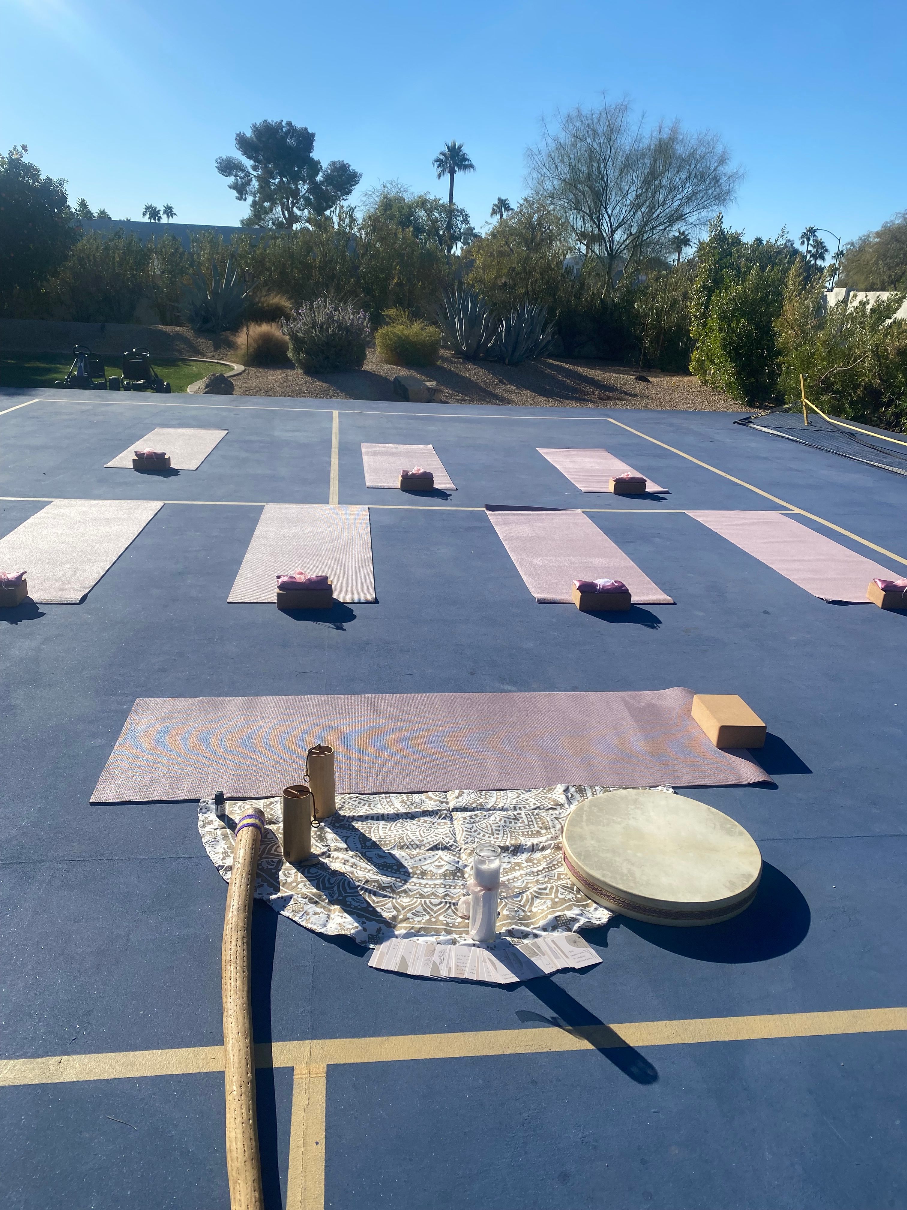 Sunny outdoor yoga setup on a blue sports court with pink mats and blocks, drum and sound instruments in foreground, desert plants and palm trees in the background