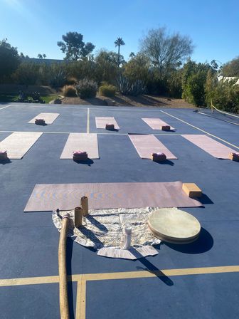 Sunny outdoor yoga setup on a blue sports court with pink mats and blocks, drum and sound instruments in foreground, desert plants and palm trees in the background