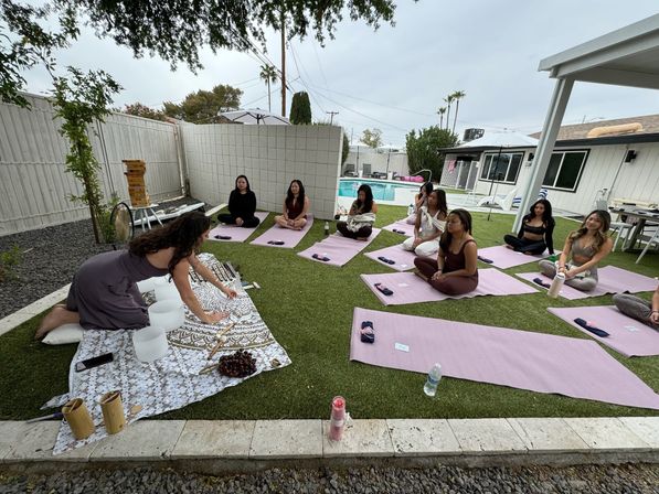 Poolside backyard sound-bath and yoga session on green lawn, instructor arranging crystal singing bowls while participants sit on pink yoga mats near a residential pool and patio.