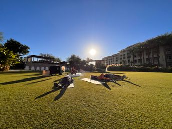 People lying on mats on a sunlit resort lawn during golden hour, palm trees and multi-story hotel balconies in the background, outdoor relaxation on manicured green grass.