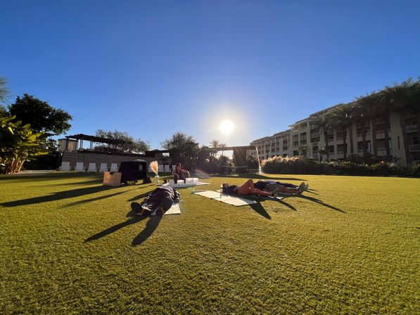 People lying on mats on a sunlit resort lawn during golden hour, palm trees and multi-story hotel balconies in the background, outdoor relaxation on manicured green grass.