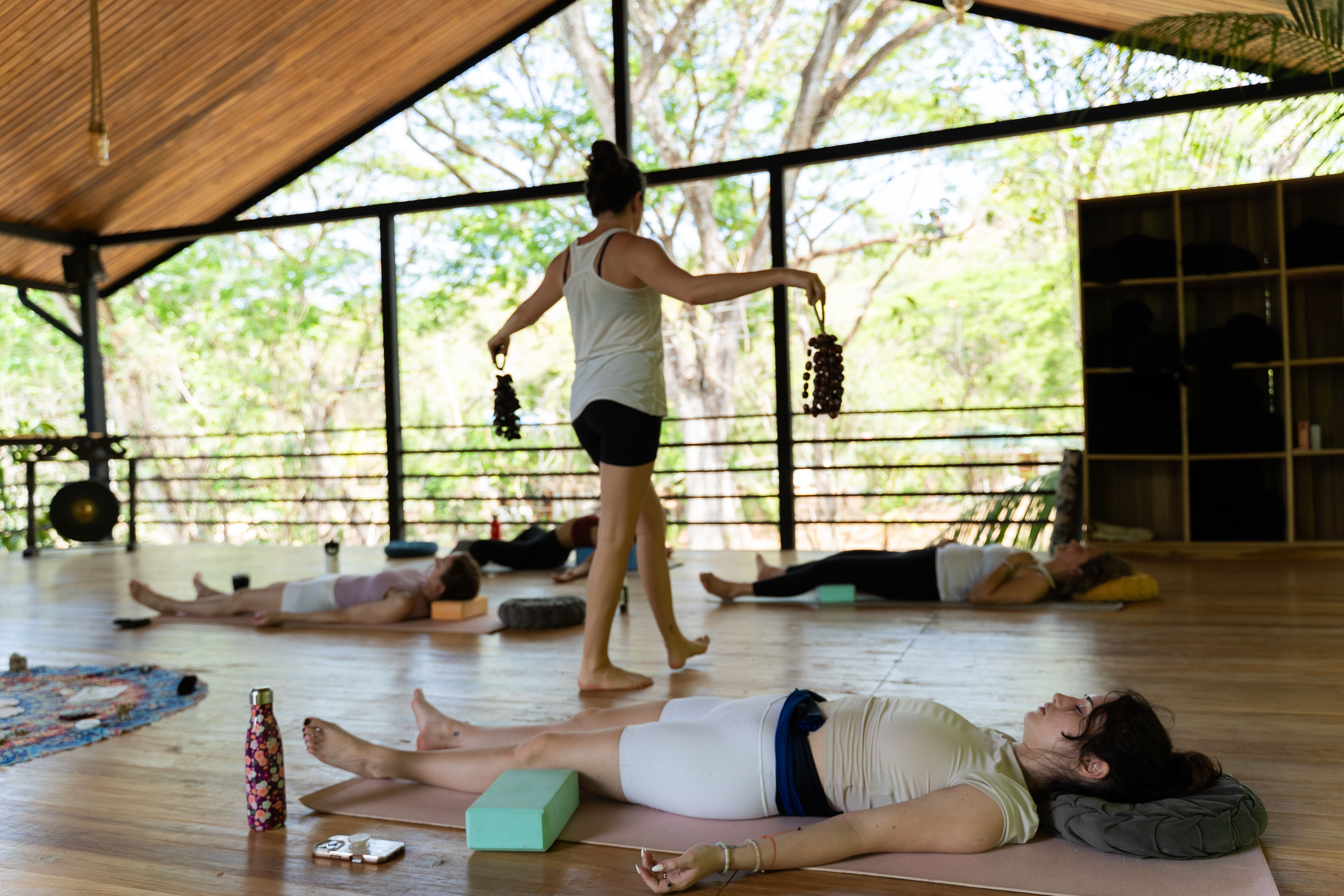 Open-air tropical yoga pavilion with students in savasana on mats while an instructor walks through holding sound-healing shakers during a relaxed wellness yoga session.