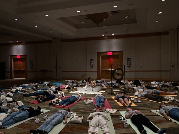 Group sound bath in a dim hotel ballroom — people lying on yoga mats around a gong and crystal singing bowls for guided meditation and relaxation.