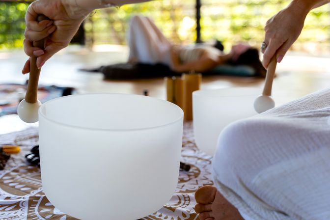 Close-up of hands playing white crystal singing bowls with mallets during a sound bath meditation in a sunlit open-air wellness retreat, a person reclining on a mat blurred in the background.