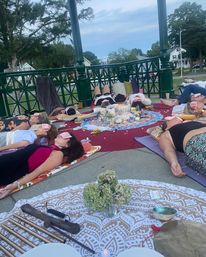 Cozy evening sound‑bath/meditation in a green gazebo at a neighborhood park — people lying on mats with eye masks around a central floral altar, candles and instruments.