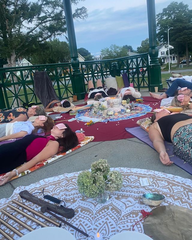 Group of people lying on mats with eye masks for an outdoor meditation/sound healing circle in a green gazebo, colorful blankets and a central arrangement of candles, flowers and instruments in a park setting.