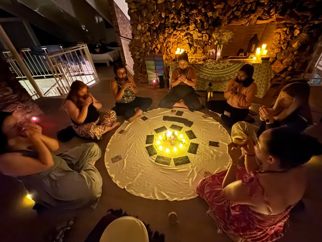 Candlelit women's circle in a cozy indoor space, participants seated cross-legged around a white cloth with glowing candles and oracle/tarot cards, stone-wall altar with more lit candles in the background