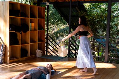 Sunlit tropical yoga retreat on an open wooden deck — a person lying relaxed on a mat while an instructor swings a handheld gong beside wooden cubby shelves and lush jungle foliage.
