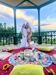 Two women smiling barefoot in a lakeside gazebo at sunset with a colorful mandala picnic setup of cushions, candles, flowers and sunflowers on a red blanket overlooking a park and water.