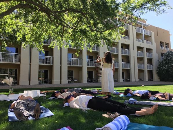 Outdoor sound-bath relaxation on a sunlit hotel lawn under a large shade tree, participants lying on yoga mats with eye masks and rolled towels while an instructor stands nearby in front of a balcony-lined building.