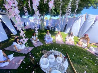 Women in white dresses and floral crowns seated on pink mats in a sunny backyard meditation and sound-bath circle beneath hanging pink flowers and white drapes, with crystal singing bowls and instruments on a patterned cloth in the center.