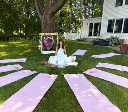 Backyard sound bath setup on green lawn with pink yoga mats radiating from a central area where a woman sits by crystal singing bowls under a large tree beside a white house.
