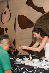 Smiling woman rings small bronze tingsha cymbals during an indoor sound-healing session over a candlelit table with bowls, a wicker chair, and abstract wall art.