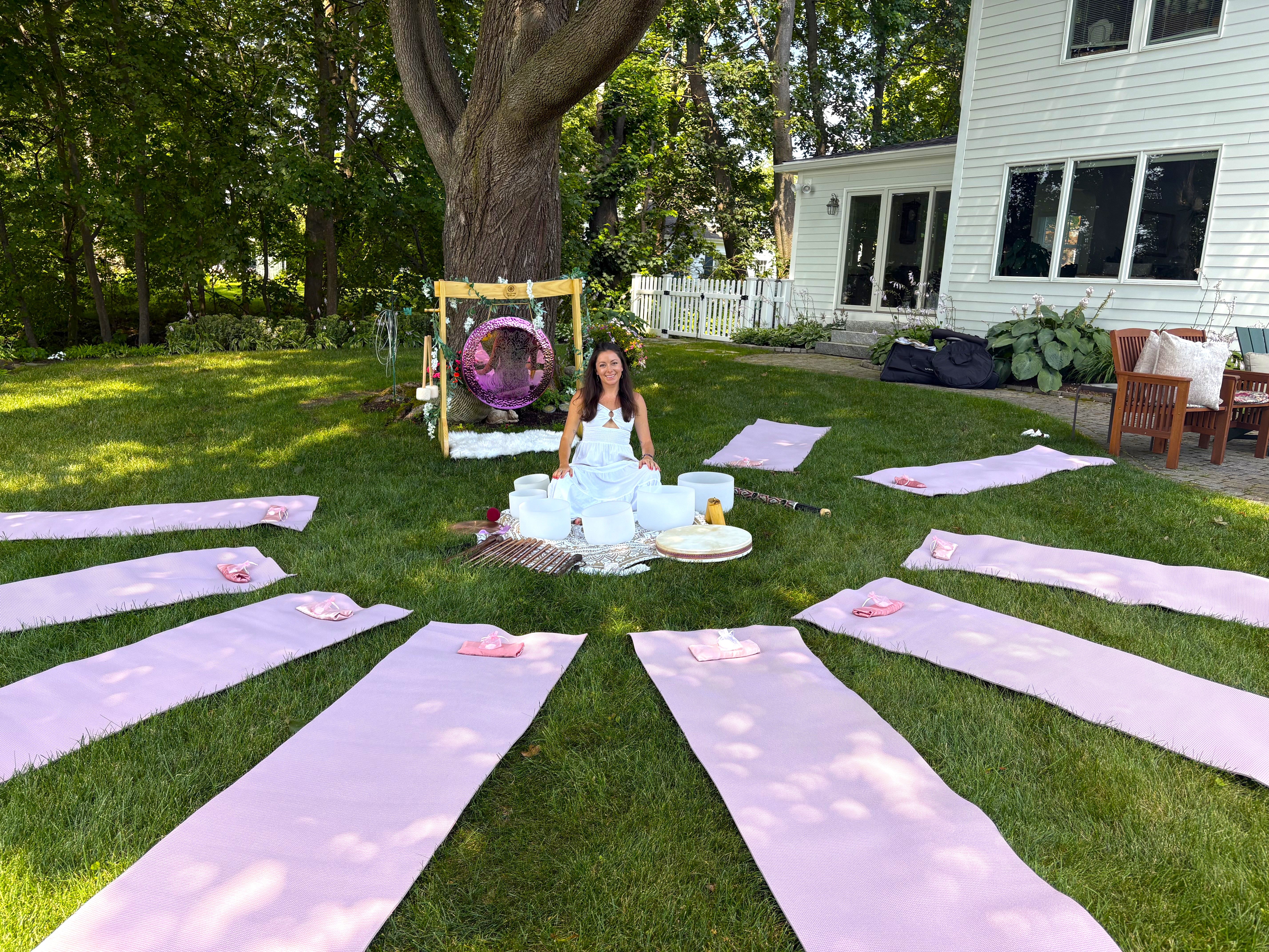 Outdoor backyard yoga and sound-healing setup with pink yoga mats arranged in a circle on green lawn, a woman seated by crystal singing bowls under a large tree, purple hanging swing and white house in the background.
