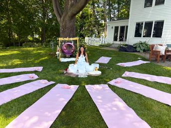 Outdoor backyard yoga and sound-healing setup with pink yoga mats arranged in a circle on green lawn, a woman seated by crystal singing bowls under a large tree, purple hanging swing and white house in the background.