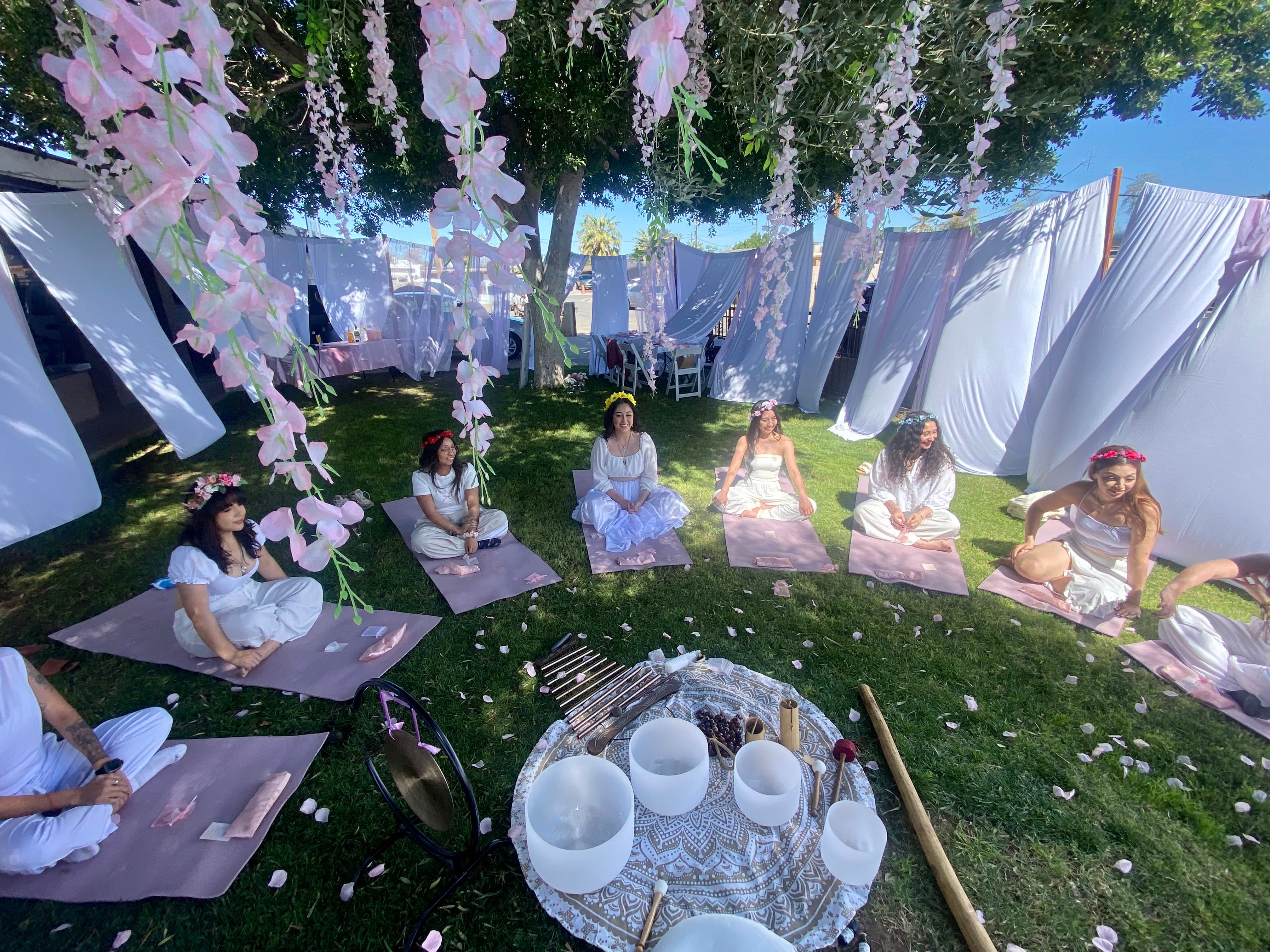 Outdoor garden meditation circle with participants in white dresses and floral crowns seated on pink mats under a canopy of hanging pink flowers and white drapes, surrounding crystal singing bowls and sound-healing instruments on a patterned cloth on the sunlit lawn.