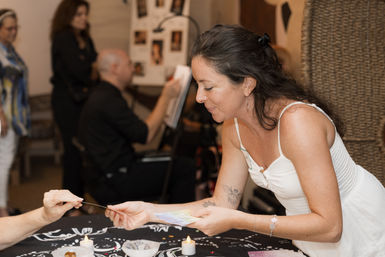 Woman in a white dress leans over a candlelit tarot table, exchanging colorful oracle cards during a lively indoor fair or community event.