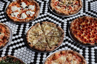 Assorted Neapolitan-style artisan pizzas with wood-charred crusts on metal trays—pepperoni, margherita, ricotta-topped and a thin-sliced white pizza—arranged on a black-and-white hexagon tile table.