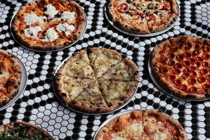 Assorted Neapolitan-style artisan pizzas with wood-charred crusts on metal trays—pepperoni, margherita, ricotta-topped and a thin-sliced white pizza—arranged on a black-and-white hexagon tile table.