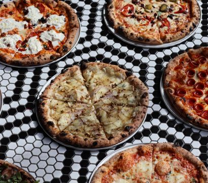 Assorted Neapolitan-style artisan pizzas with wood-charred crusts on metal trays—pepperoni, margherita, ricotta-topped and a thin-sliced white pizza—arranged on a black-and-white hexagon tile table.